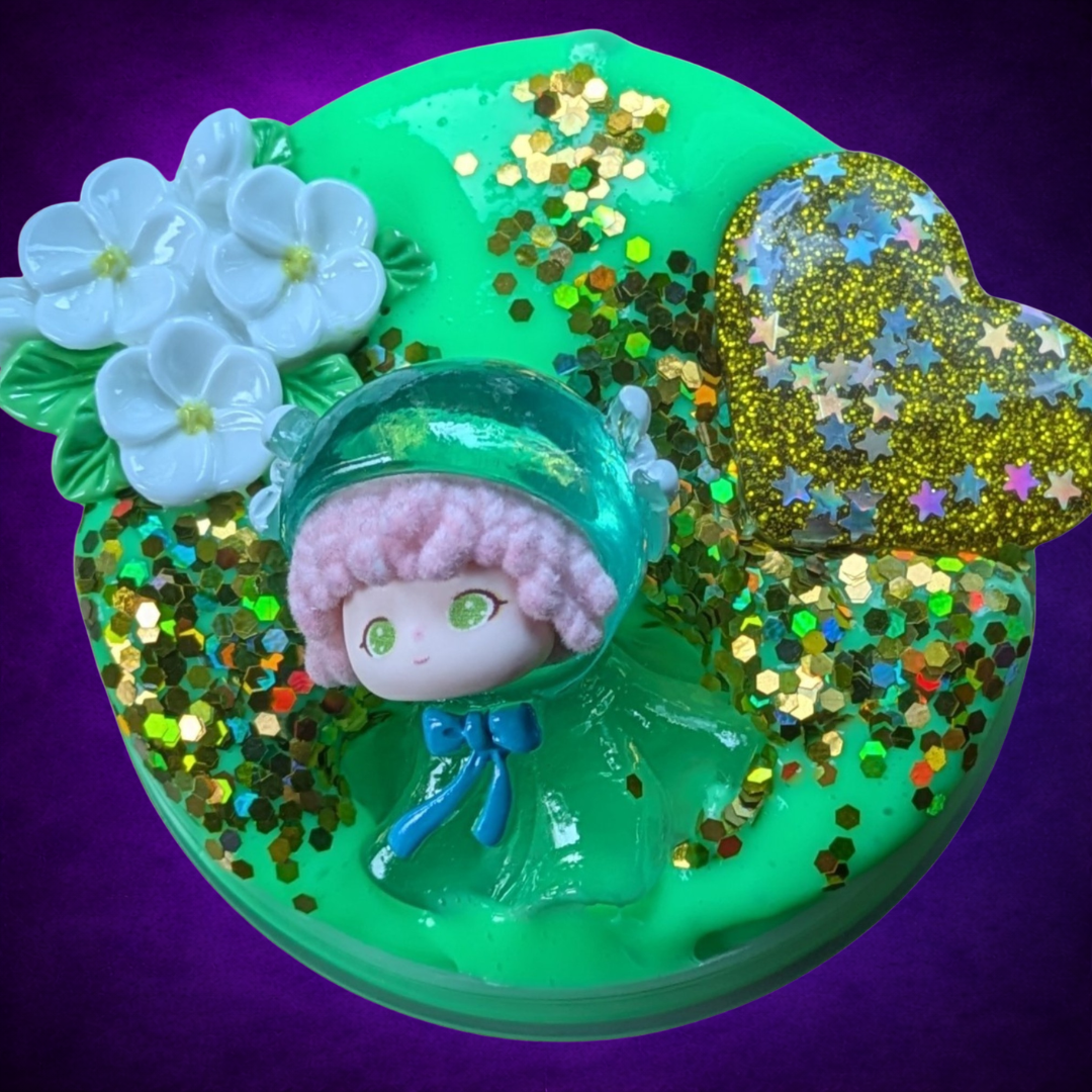 Overhead view of Ériu’s Promise green slime with gold glitter, a gold heart, white flowers and figure.